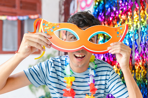Young Brazilian man having fun with a carnival mask, set against the backdrop of a house decorated for a carnival party. Perfect for capturing the joy of Carnival celebrations.