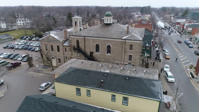 A drone captures an aerial view of Niagara-on-the-Lake's downtown courthouse, on a quaint old street filled with shops, as light snow falls in winter.