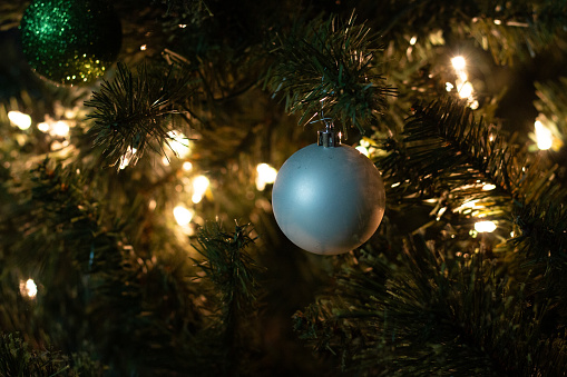 A white, spherical ornament hangs in the center of the frame, on a green Christmas tree. The tree is strung with warm white lights. There is a glittery, green ornament in the top right corner.