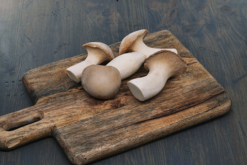 Fresh eryngi mushrooms, vary in sizes, rest on a wooden cutting board, background grey-blue toned wooden surface, vibrant scene of food preparation