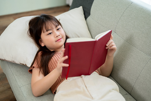 Young happy and cute Asian girl is reading a book while lying on a sofa in the living room. Kid, hobby, leisure, education
