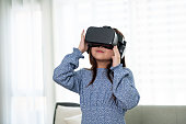 A cute young Asian girl is wearing VR headset and experiencing virtual reality game at home.