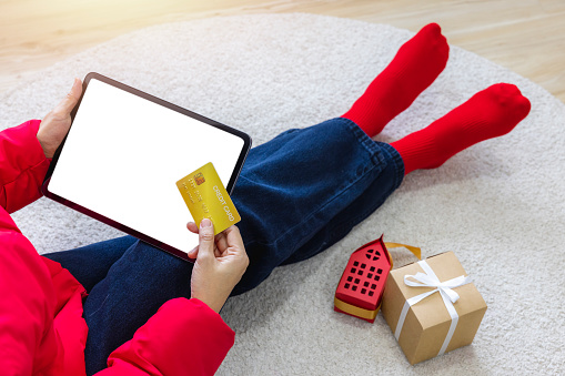 A woman shopping online sitting on carpet at home with a tablet with a white screen isolated using a gold credit card. Use your credit card to tap the screen to verify your identity. Clipping Path.