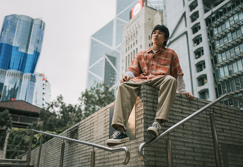 Asian Chinese young man sitting on wall with Hong Kong city background looking away with cool attitude