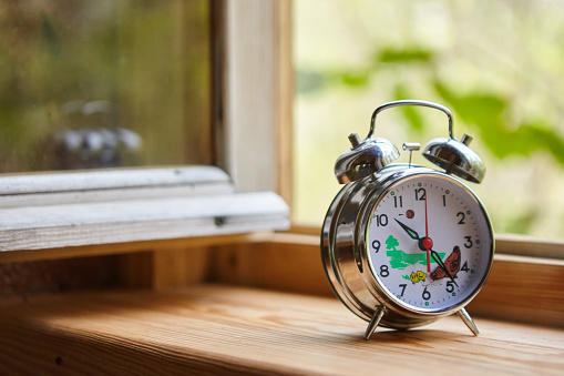 A mechanical alarm clock stands on the windowsill near an open window. The time of year is spring or summer.