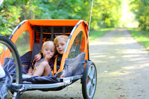 Two young children are sitting together in a pull behind bicycle trailer as they ride down a bike trail in the woods