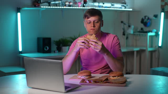 Portrait of hungry young man eating taco burger and texting on mobile phone, sitting at table with laptop and burgers during watching online movie