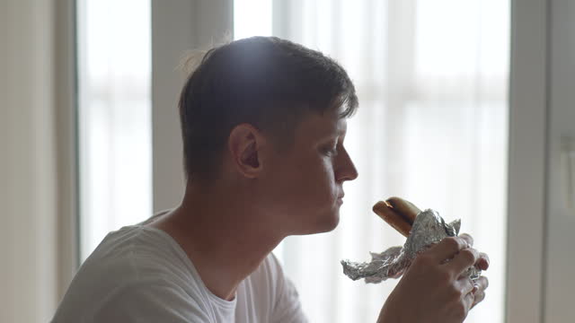 Close-up side view of hungry man eating delicious sandwich at home on background of window. Handsome male enjoying vegetarian dinner indoors.