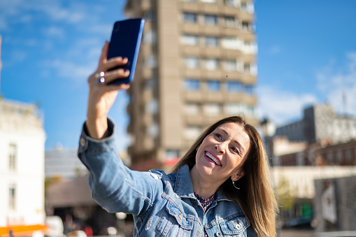 Smiling Beautiful Woman in Casual Clothes. Happy and Smiling Female Taking Selfie Self Portrait of Himself on Smartphone Camera. Having Fun on the Street Background.