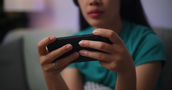 Portrait close up of Young Asian woman wearing glasses and headphones enjoys playing online esport games on smartphone sitting on sofa in the living room at home,Gamer lifestyle concept.