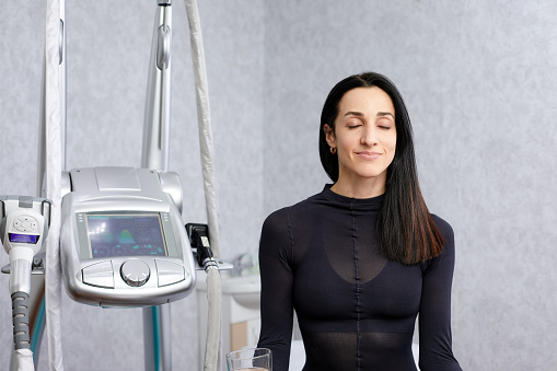 woman in a black suit for LPG massage sits with closed eyes before LPG massage. Anti-cellulite body care concept. LPG massage machine on background