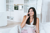 beautiful fashion model with Asian roots is sitting on couch at home talking on phone looking away smiling enjoying communication with a friend. A young American woman spends time at home on weekends