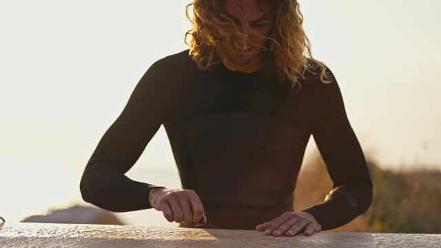 A guy with curly hair in a wetsuit sitting processes his Surf and cleans it on the beach in the morning. Preparing for surf swimming
