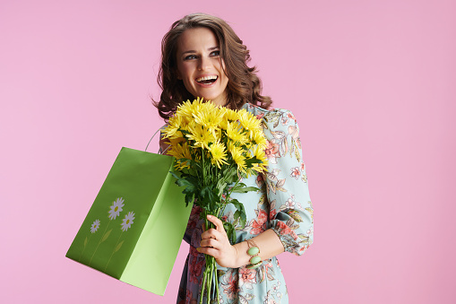 happy trendy female with long wavy brunette hair with yellow chrysanthemums flowers and green shopping bag against pink background.