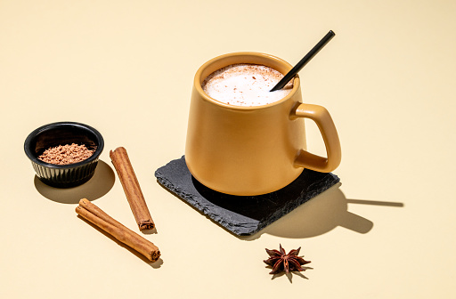 Yellow cup  with foamy drink coffee latte, hot chocolate or cocoa with cinnamon sticks and anise star on a yellow background with hard shadow. Free space for text.