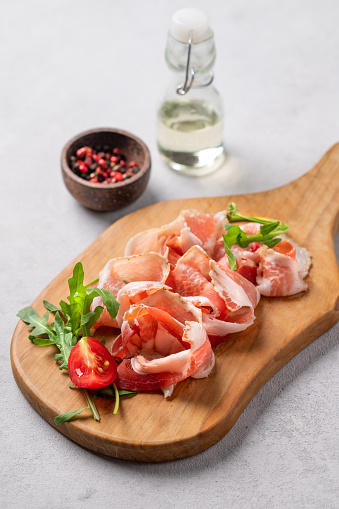 Snack wooden board with bacon or prosciutto on a light background with spices, tomatoes and herbs. Antipasto concept.