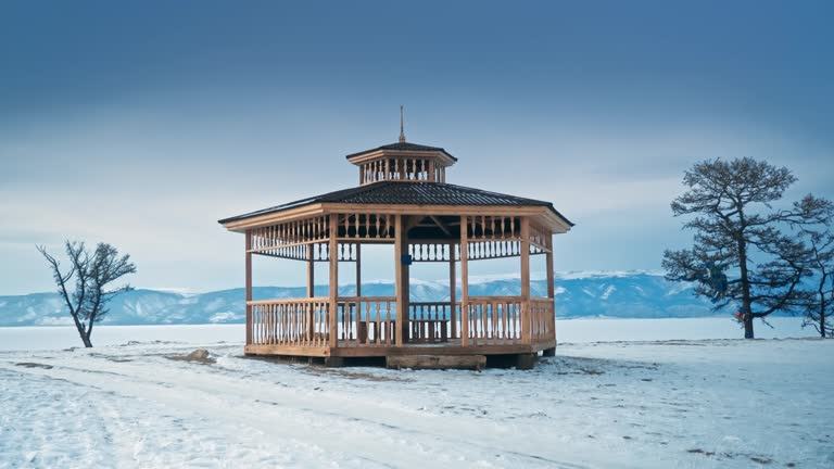 Empty wooden gazebo with benches, balusters, pitched roof and belvedere surrounded by trees with fallen leaves in winter on the shore of Lake Baikal against the background of a mountain range, clear weather. Siberia, Russia, Olkhon.