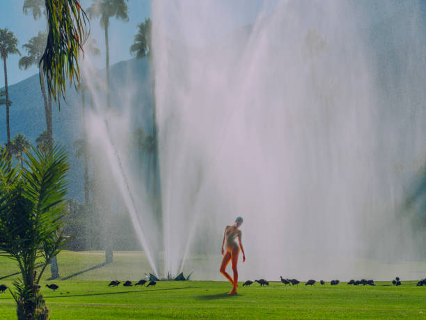 The photo is focused on the fountain in the front and middle of the shot that shoots up and curls over at the top. The fountain is encased in two tan tiled sides of the pool with the main part of the pool in the middle of the shot and the hot tub cornered and sectioned off at the top of the shot.