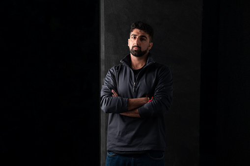 A studio portrait of a young adult male standing with his arms crossed. He is standing against a black background, the lighting is low and dramatic. He has a beard and is wearing a black quarter zip pullover, with his head cocked to one side whilst looking into camera with a neutral expression. On his wrist he wears a metal Kara bracelet signifying his religion.