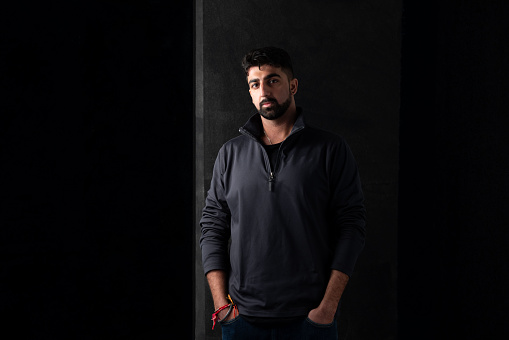 A studio portrait of a young adult male standing with his hands in his pockets. He is standing against a black background, the lighting is low and dramatic. He has a beard and is wearing a black quarter zip pullover, with his head cocked to one side whilst looking into camera with a neutral expression. On his wrist he wears a metal Kara bracelet signifying his religion.