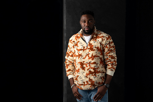 A studio portrait of a young adult male standing with his hands in his pockets. He is standing against a black background, the lighting is low and dramatic. He has a beard and is wearing a funky patterned pullover fleece, looking into camera with a neutral expression.