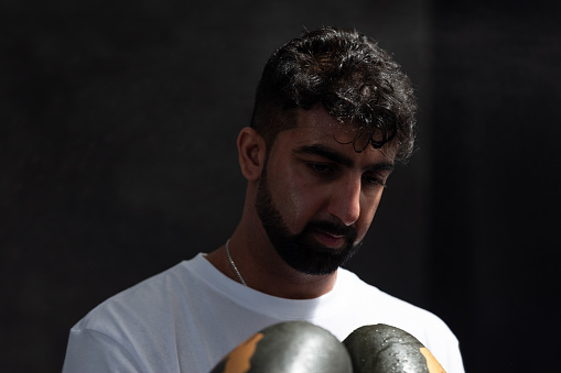 A cropped studio shot of a young adult male wearing boxing gloves, holding his hands in front of his chest, partly off-frame, whilst looking down. He is standing against a black background, the lighting is low and dramatic. He has a beard and is wearing a white t-shirt, with a serious expression and moisture on his hair and skin.