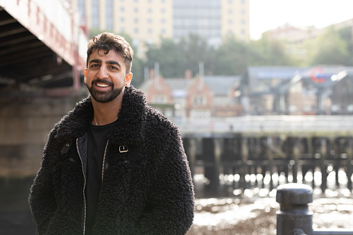 A waist-up shot of a young adult male leaning against a railing beside a river in the city centre of Newcastle upon Tyne, North East England. He has a beard and dark hair, looking directly at the camera and smiling. He is wearing casual clothing and behind him is the river and tall buildings which are out of focus.