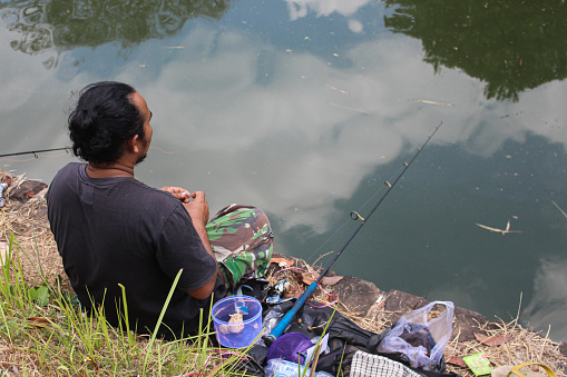 Banjar City, Indonesia - Oct 1, 2023: Participants in sport fishing tournaments. Asian man in sportswear with fishing pole is fishing by the lake and waiting for his bait to be struck on a sunny day.