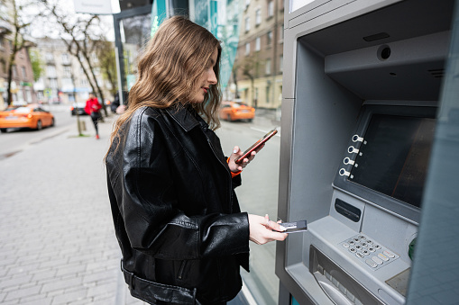 Young woman in leather jacket inserting a credit card to ATM outdoor, while looking on smartphone.