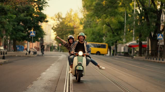 Happy couple guy with long curly hair in a plaid shirt and denim jacket rides with his happy brunette girlfriend in a plaid shirt on a moped, the girl puts her arms and legs forward having fun and rejoicing while riding along a wide street with her