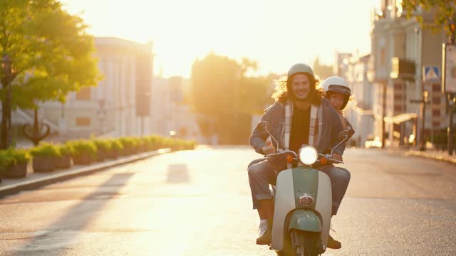 Happy couple guy with long curly hair in a green motorcycle helmet and denim jacket rides with his brunette girlfriend who looks out from behind his shoulder while riding a green moped along a beautiful summer city street on a golden sunrise in the summer