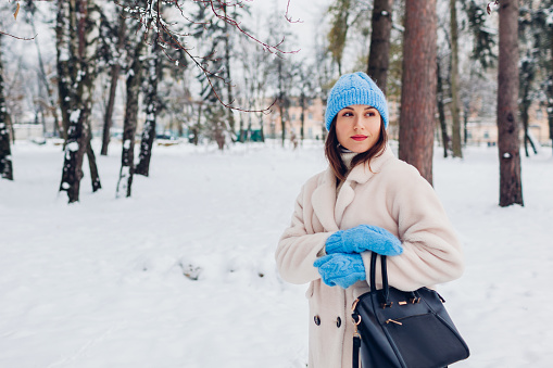 Stylish young woman walking in snowy winter park wearing white sheepskin fur coat, blue hat, mittens, holding handbag. Warm clothes. Space