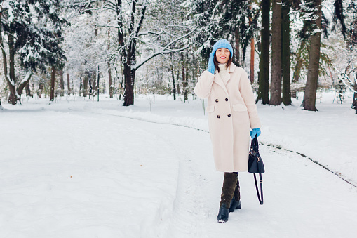 Full body portrait of young woman walking in snowy winter park wearing stylish white fur coat, holding handbag. Warm clothes for cold weather. Space