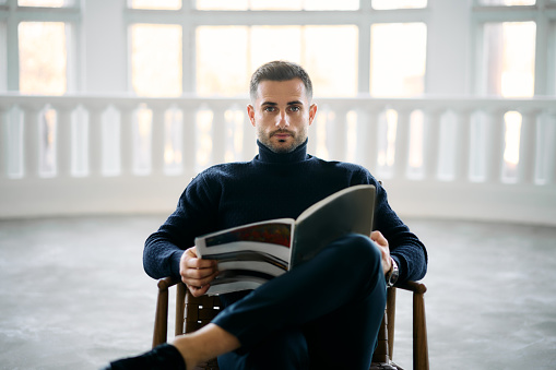 Young handsome man reading magazine seated comfortably in chair in modern white room. Relaxing concept