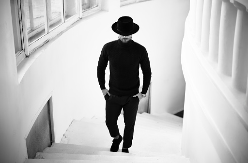 Stylish man in black clothing outfit and hat, looking downwards walking on marble staircase. Black and white photo