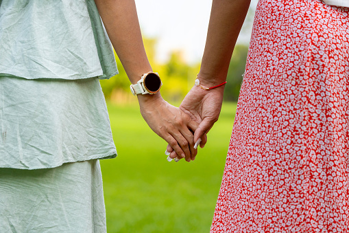 Diversity of LGBTQ+ lesbian couple is relaxingly holding their hand while walking together in public park during summer season to enjoy the leisure time for coming out of closet and inclusion