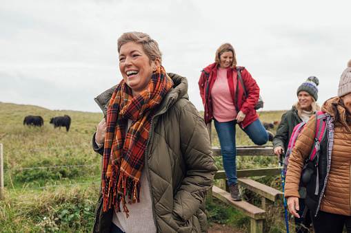 Four mature women wearing warm, casual, outdoor clothing and accessories on a day out in Northumberland. They smile and enjoy the views as they walk through the British countryside. One of the women has short blond hair as she is in recovery after beating breast cancer.