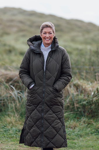 A mature woman wearing warm, casual, outdoor clothing and accessories on a day out in Northumberland. She is smiling and looking at the camera as she stands amongst sand dunes. She has short blond hair as she is in recovery after beating breast cancer.