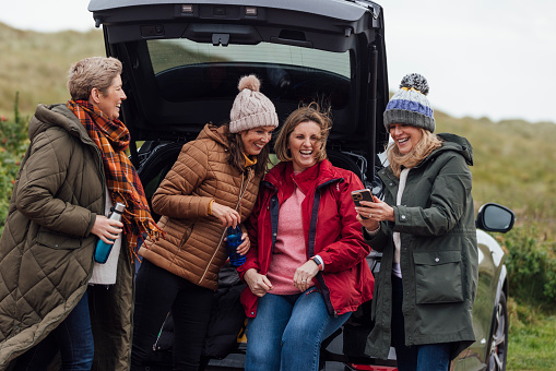 Four mature women wearing warm, casual, outdoor clothing and accessories on a day out in Northumberland. They stand at the trunk of an electric car and get prepared for their day ahead by the sea.