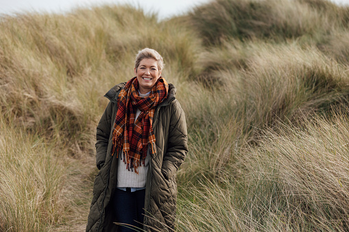 A mature woman wearing warm, casual, outdoor clothing and accessories on a day out in Northumberland. She is smiling and looking at the camera as she stands amongst sand dunes. She has short blond hair as she is in recovery after beating breast cancer.