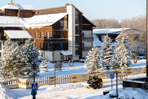 Snow-covered landscape with wooden buildings, icicles hanging from rooftops, evergreen trees laden with snow, a clear blue sky, and a small vehicle clearing the road.