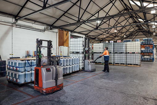 Wide shot of a blue-collar worker wearing casual clothing and reflective vests in an ink factory in Hexham, Northeastern England. He is using an electric pallet jack to organize containers filled with ink.