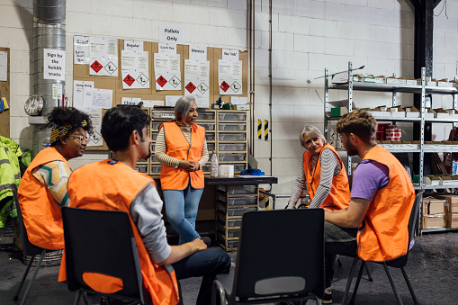 Over-the-shoulder shot of a team of five colleagues wearing casual clothing and reflective vests in a family ran ink factory in Hexham, Northeastern England. They are sat in a circle having a meeting with each other.