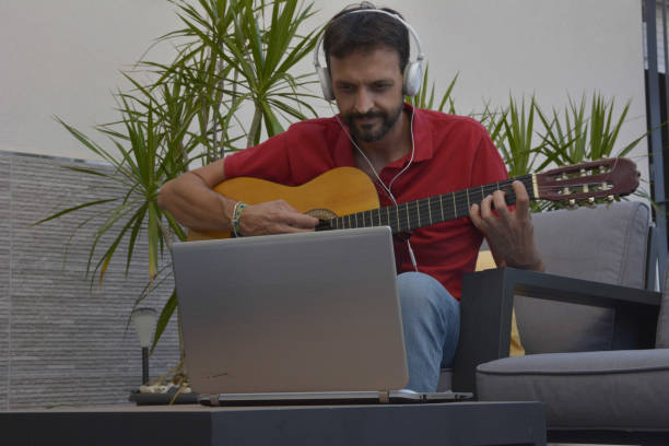 istock man doing an online course to learn to play classical guitar. He is sitting on sofa playing guitar in front of laptop while listening to lessons with helmet 1837378058