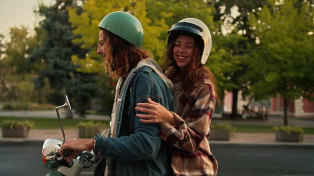 A happy brunette guy with curly hair in a denim jacket drives up on his moped, his girlfriend in a checkered shirt and a white helmet runs up to him, gets on the moped with the guy and they leave