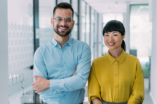 Shot of two confident business persons standing side by side in the office corridor and looking at camera. Portrait of successful businessman and businesswoman posing together in their office.