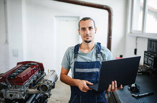 Auto mechanic using laptop while examining car engine in a workshop