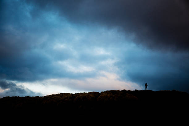 Storm clouds cover the sky as a silhouetted man stands on a hilltop.