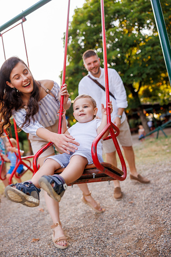 Cheerful young mother and father having fun playing with their child on playground in the park, swinging him on a swing