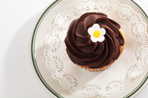 Subject: Horizontal overhead, directly above view of a cupcake frosted with swirled chocolate frosting and decorated with a white and yellow candy flower. The cupcake is on an old-fashioned diner plate with a doily—a tempting decadent treat for those who indulge in sweets and love cakes with lots of frosting.